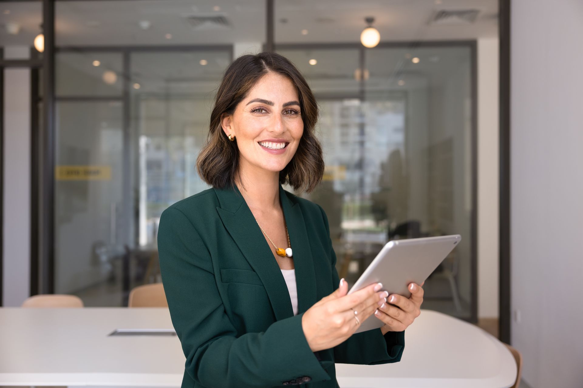 Woman in a green blazer smiling while holding a tablet in an office lobby
