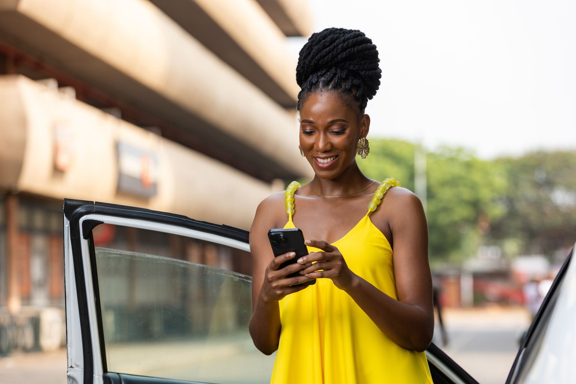 Woman in a yellow dress smiling at her phone beside an open car door.