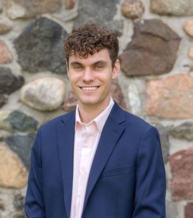 Young man with curly hair smiles, wearing a blue blazer and pink shirt against a stone wall.