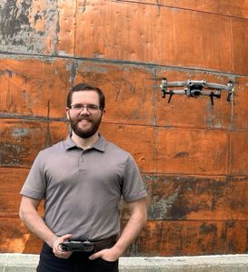 Man with drone controller, drone hovering near a rusty orange wall. He smiles, wearing glasses and a gray shirt.