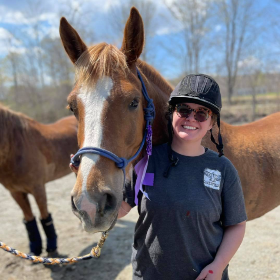 Woman in riding helmet smiles next to a brown horse with a white blaze, outdoors.