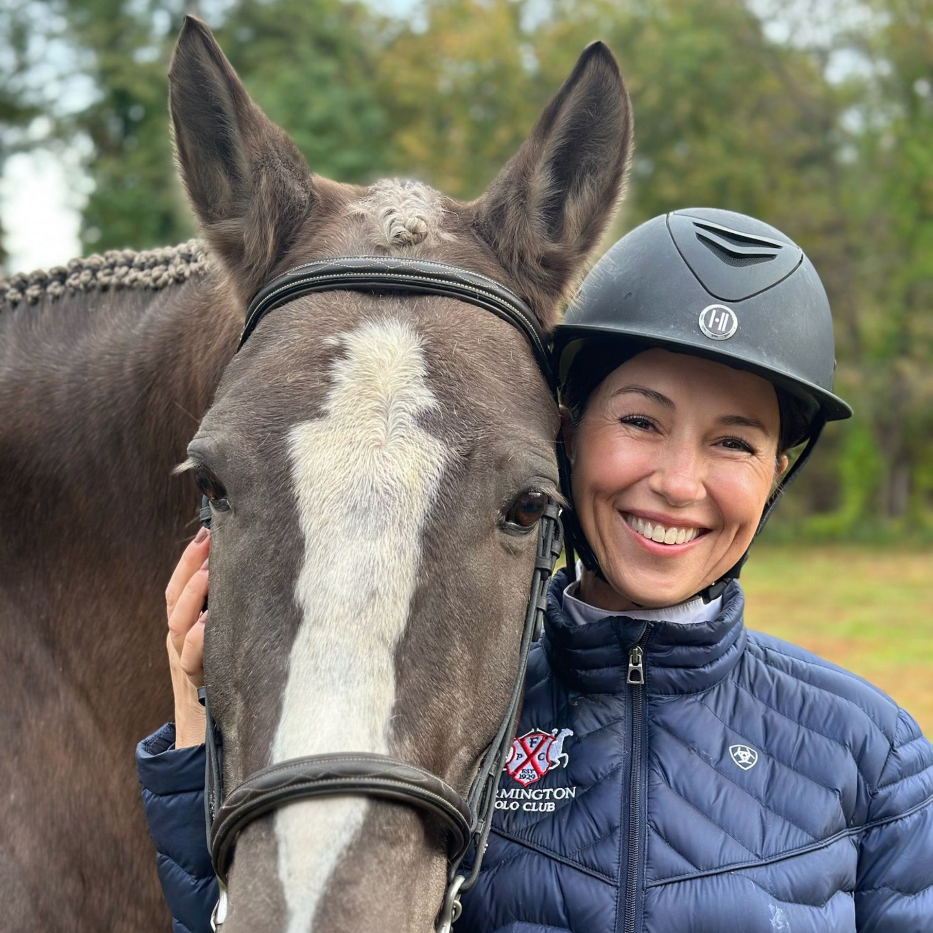 Woman in riding helmet smiles, petting a gray horse with a white face marking, outdoors.