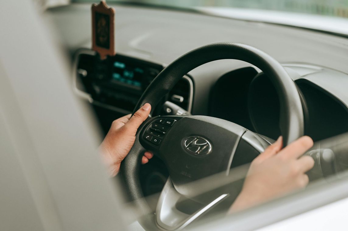 Hands on a steering wheel inside a car interior with a hanging ornament and dashboard display