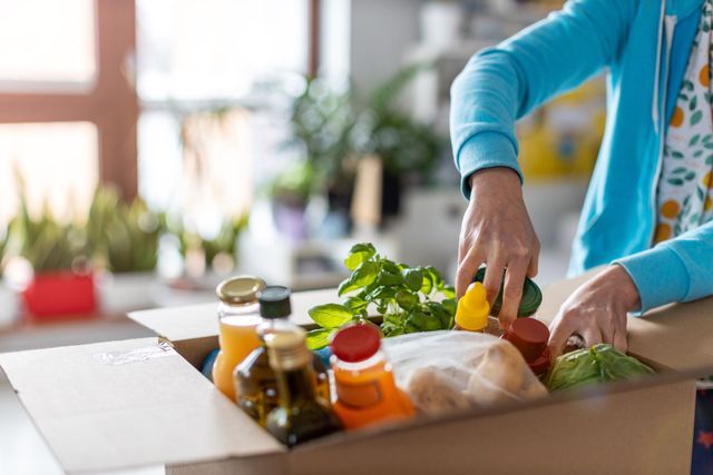 Hands unpacking groceries from a cardboard box on a countertop, near a window.