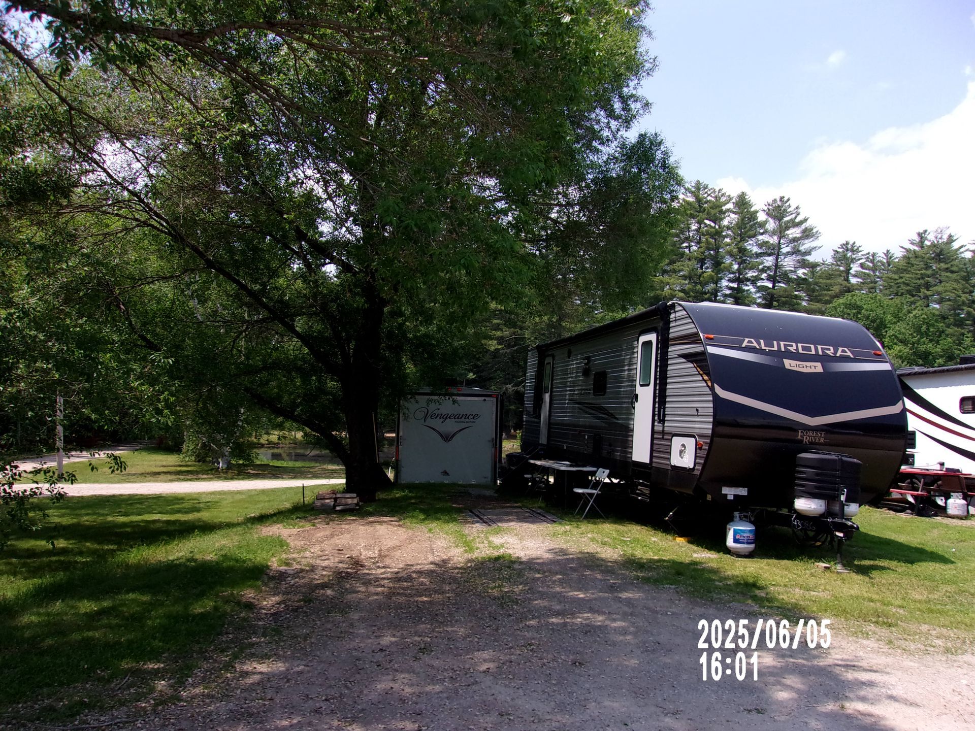 Camouflaged RV parked on a grassy campsite under trees, with a small shed nearby.