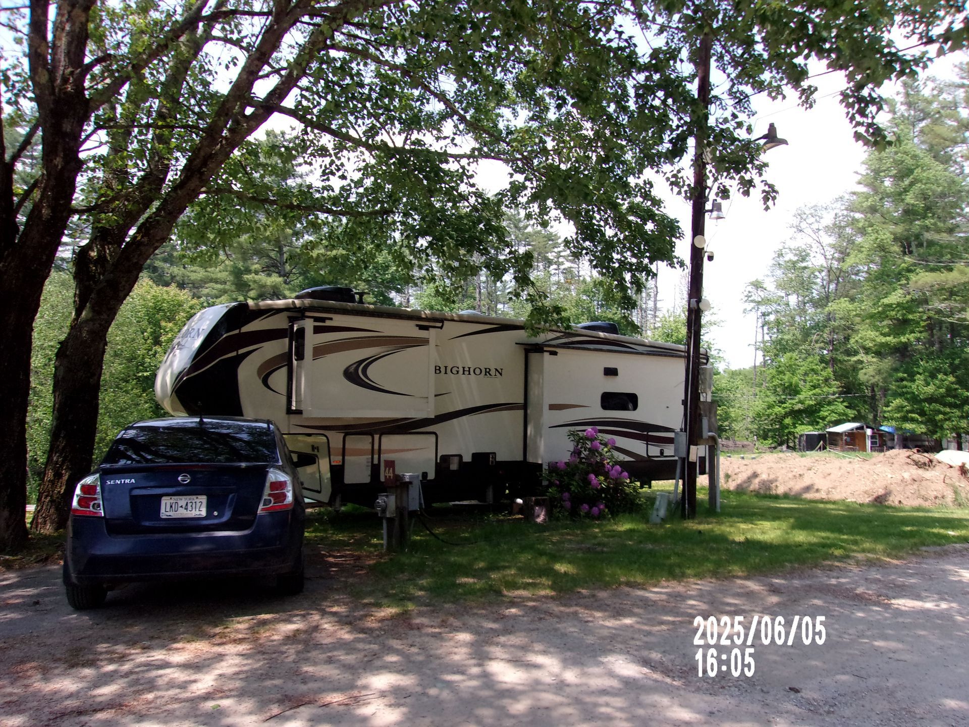 Blue SUV parked beside a tan fifth-wheel RV under trees at a campground