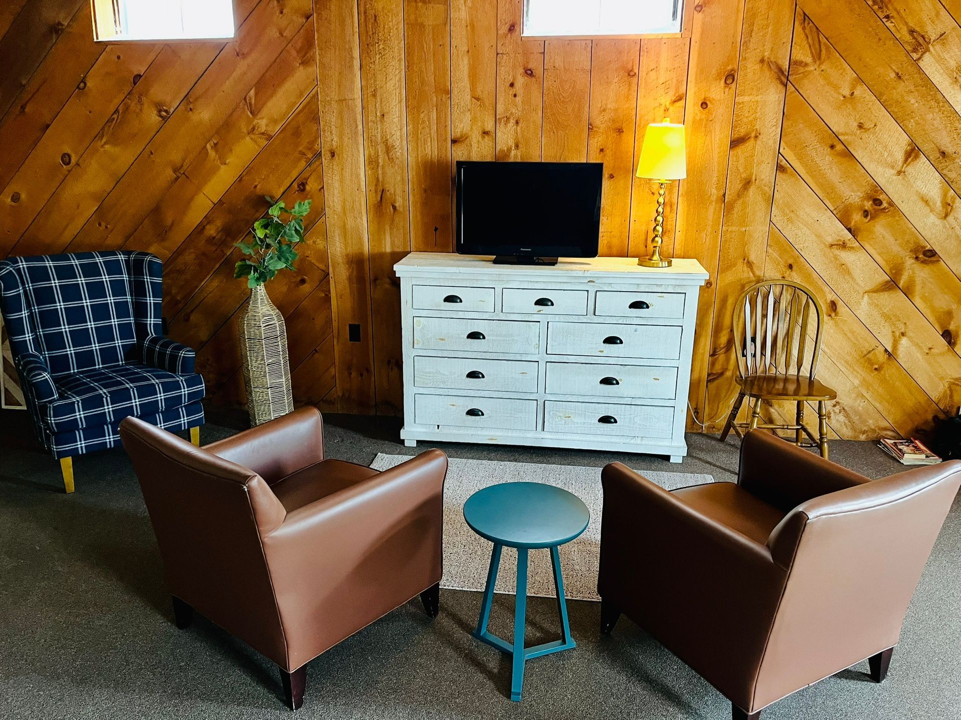 Cozy rustic sitting room with four chairs, small round table, white dresser, lamp, and TV against wood-paneled walls