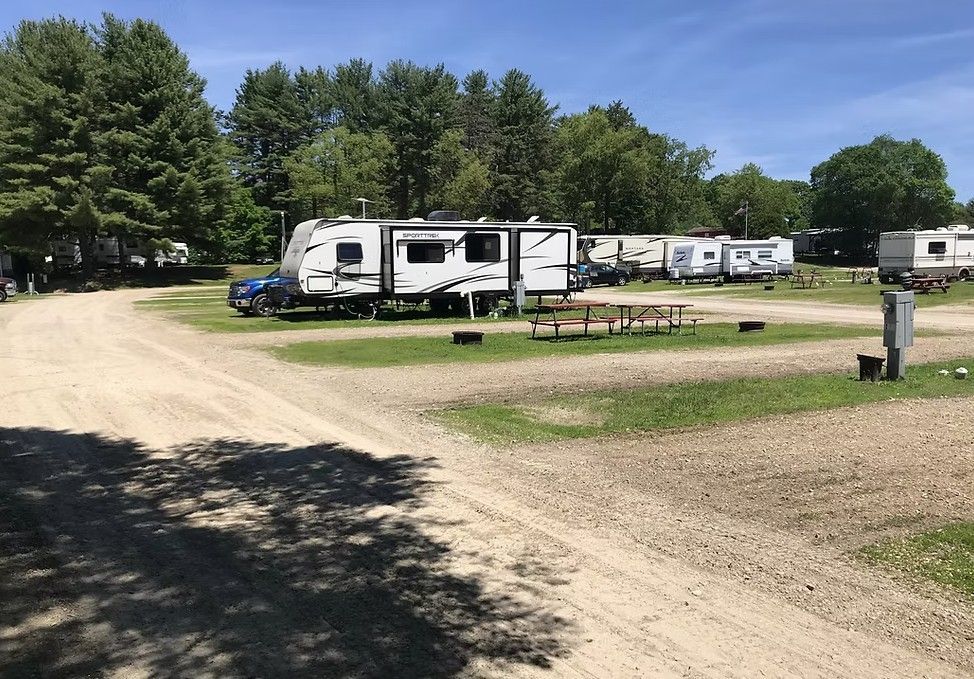 RVs parked at a gravel campground under a sunny blue sky, with trees and a dirt road.