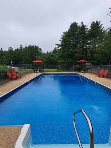 Outdoor swimming pool with blue water, red chairs, and red umbrellas surrounded by trees under an overcast sky