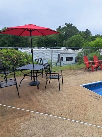 Patio with red umbrella, black table and chairs beside a blue pool and red chairs overlooking a lake