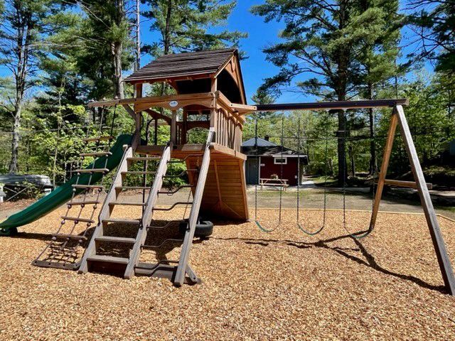 Wooden playground climbing structure and swing set on wood chips, surrounded by tall pine trees.