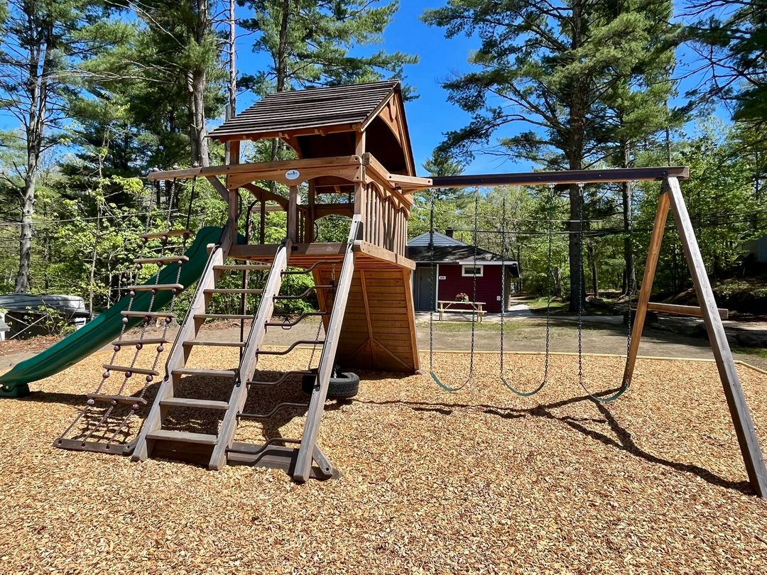 Wooden playground with slides, climbing steps, and swing set on a woodchip surface among pine trees.