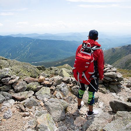 Hiker in red jacket and backpack standing on rocky mountain summit overlooking distant blue ridges