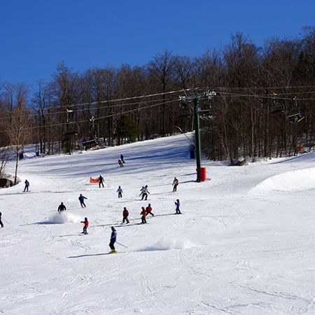 Skiers descending a snowy slope at a ski resort with a chairlift and trees under a clear blue sky