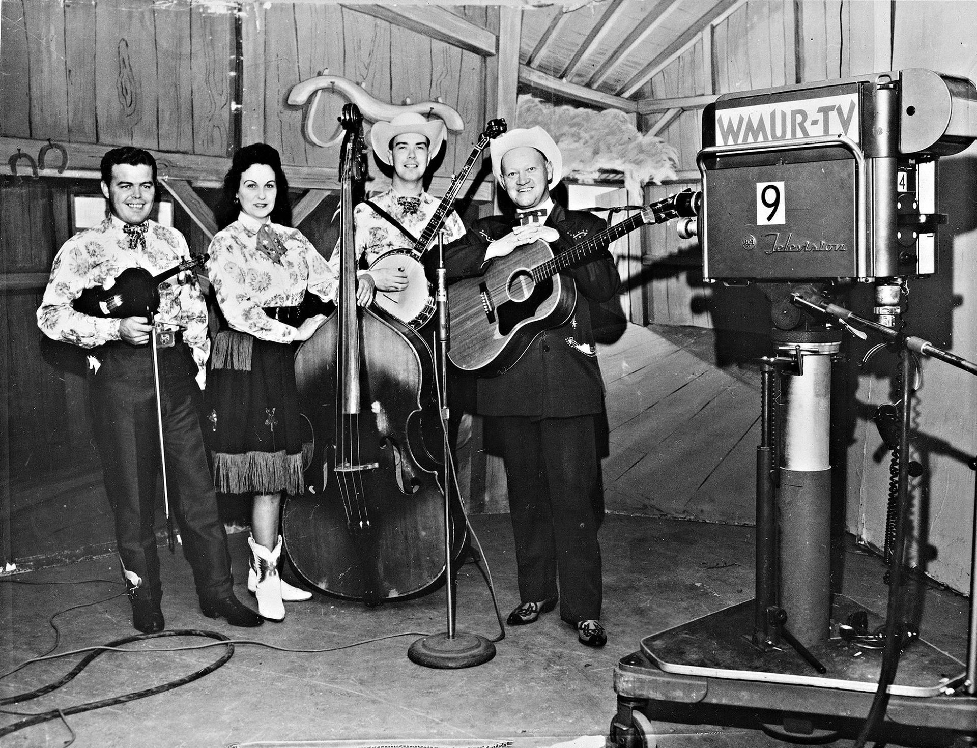 Black-and-white TV studio scene with four musicians performing beside a large camera.