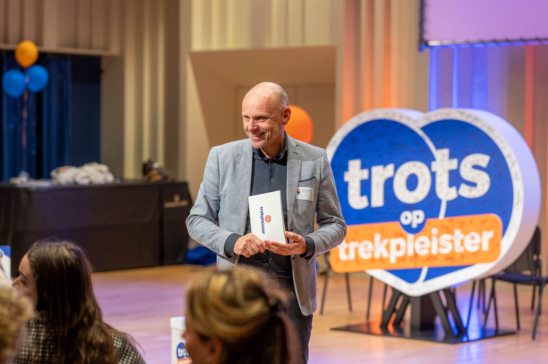 Een man met een prijs in zijn handen lacht tijdens een evenement, voor een blauw-oranje bord.