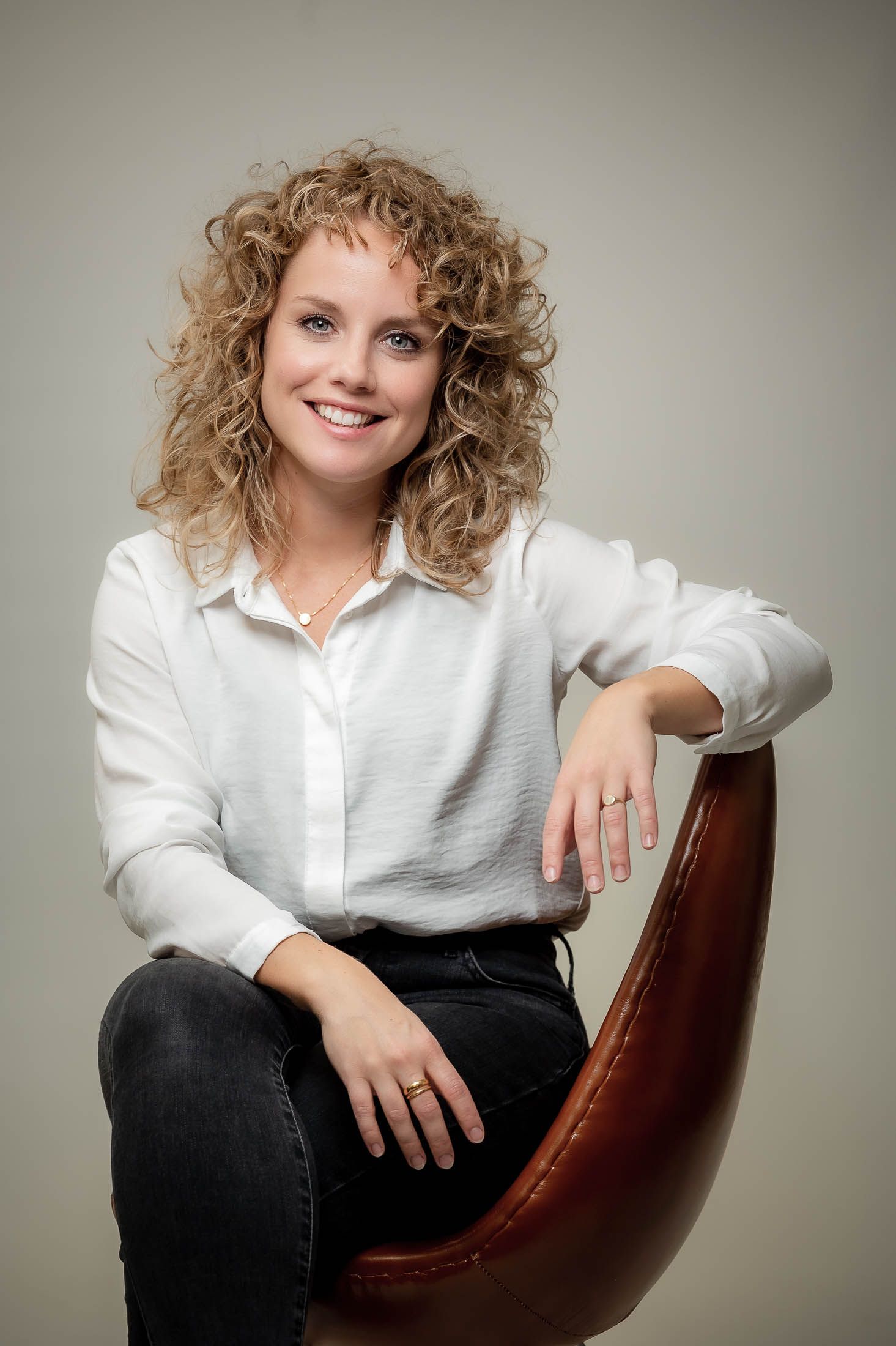 Een vrouw met krullend haar zit op een stoel en glimlacht.