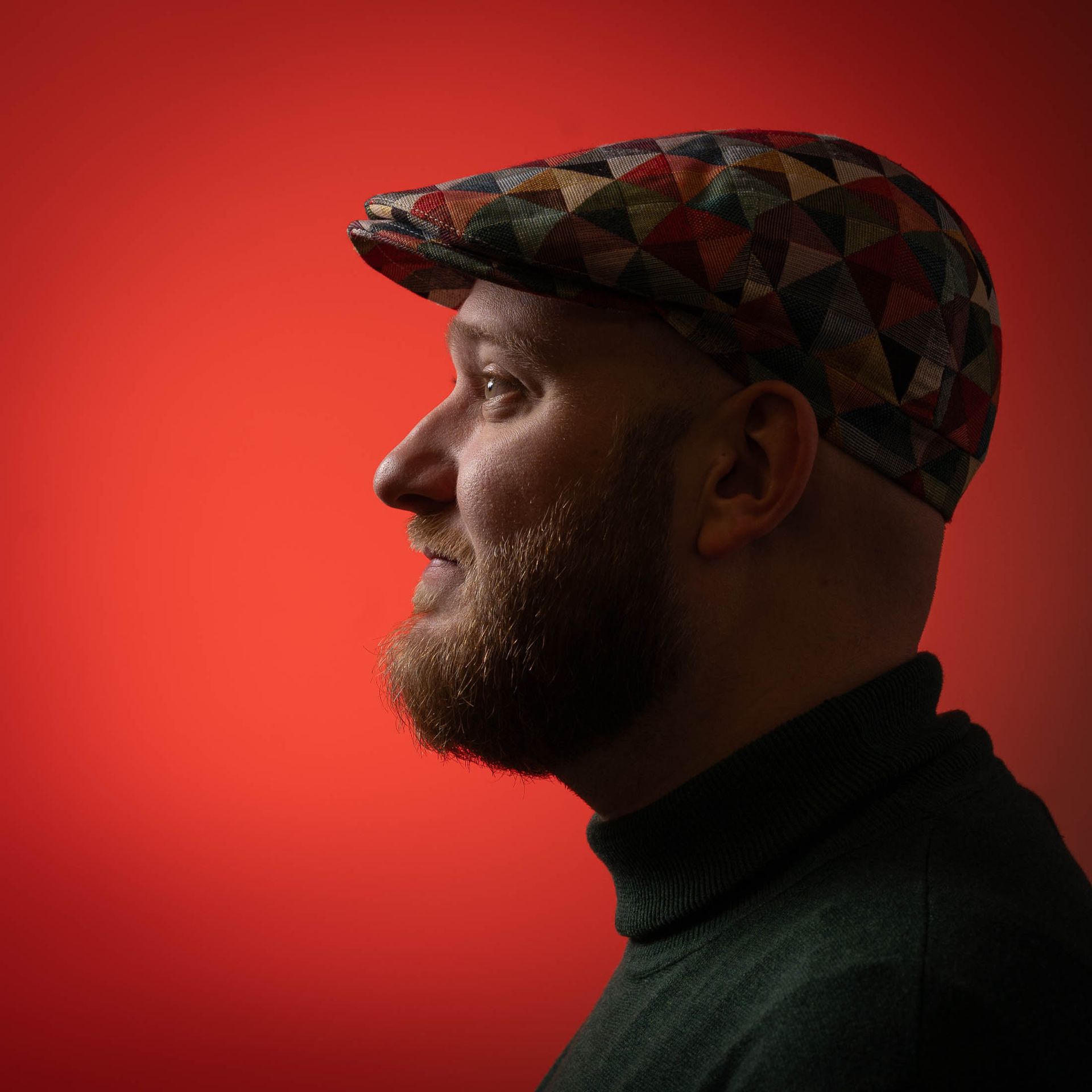 Een man met een baard draagt een platte pet en een groene coltrui.