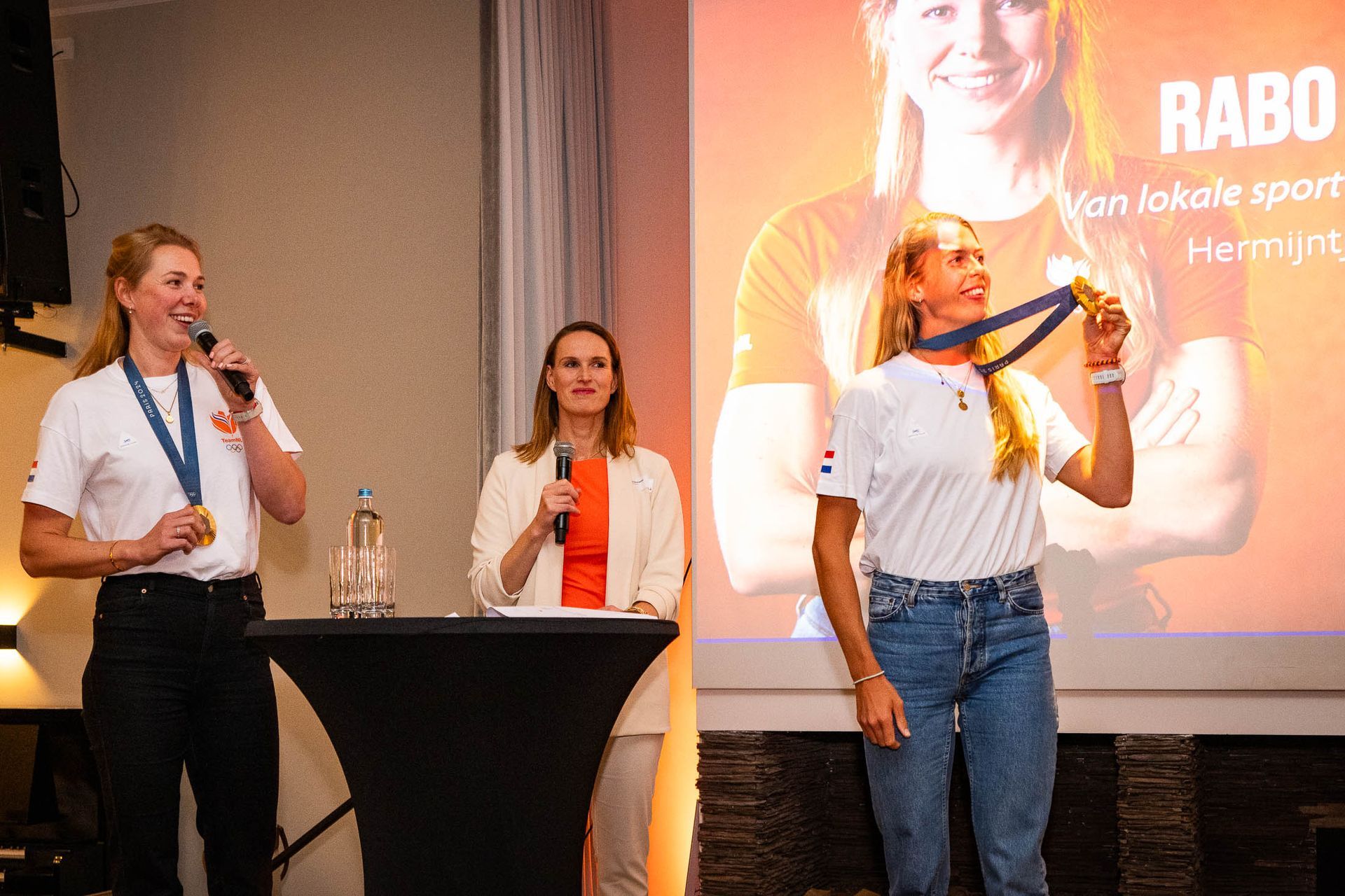 Drie vrouwen staan voor een projectiescherm en houden medailles vast.