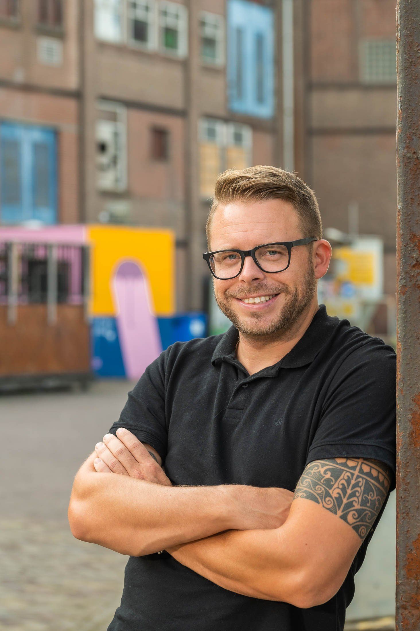Een man met een bril en een zwart shirt staat met zijn armen over elkaar voor een gebouw.