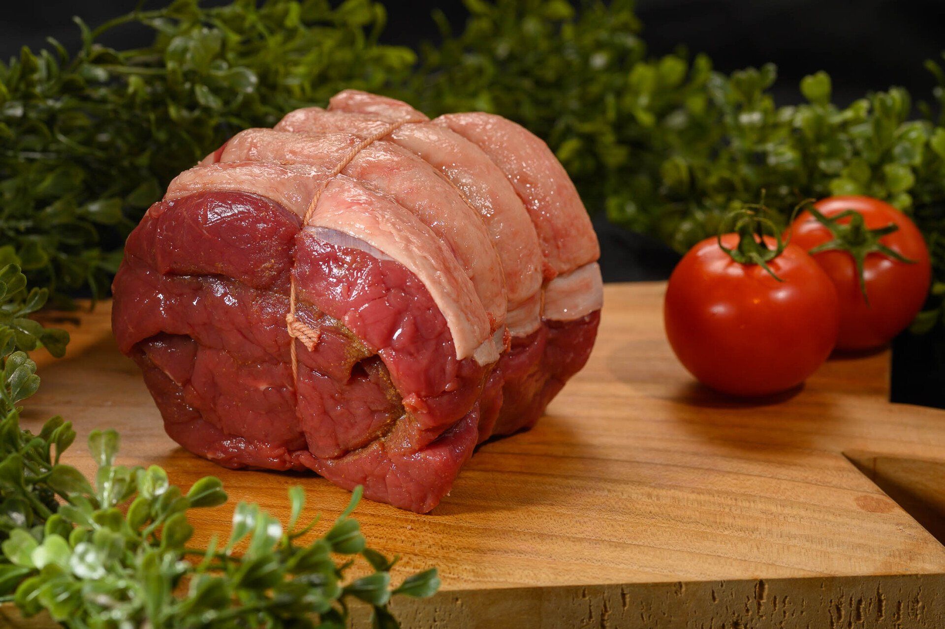 Een stuk rauw vlees ligt op een houten snijplank naast tomaten.