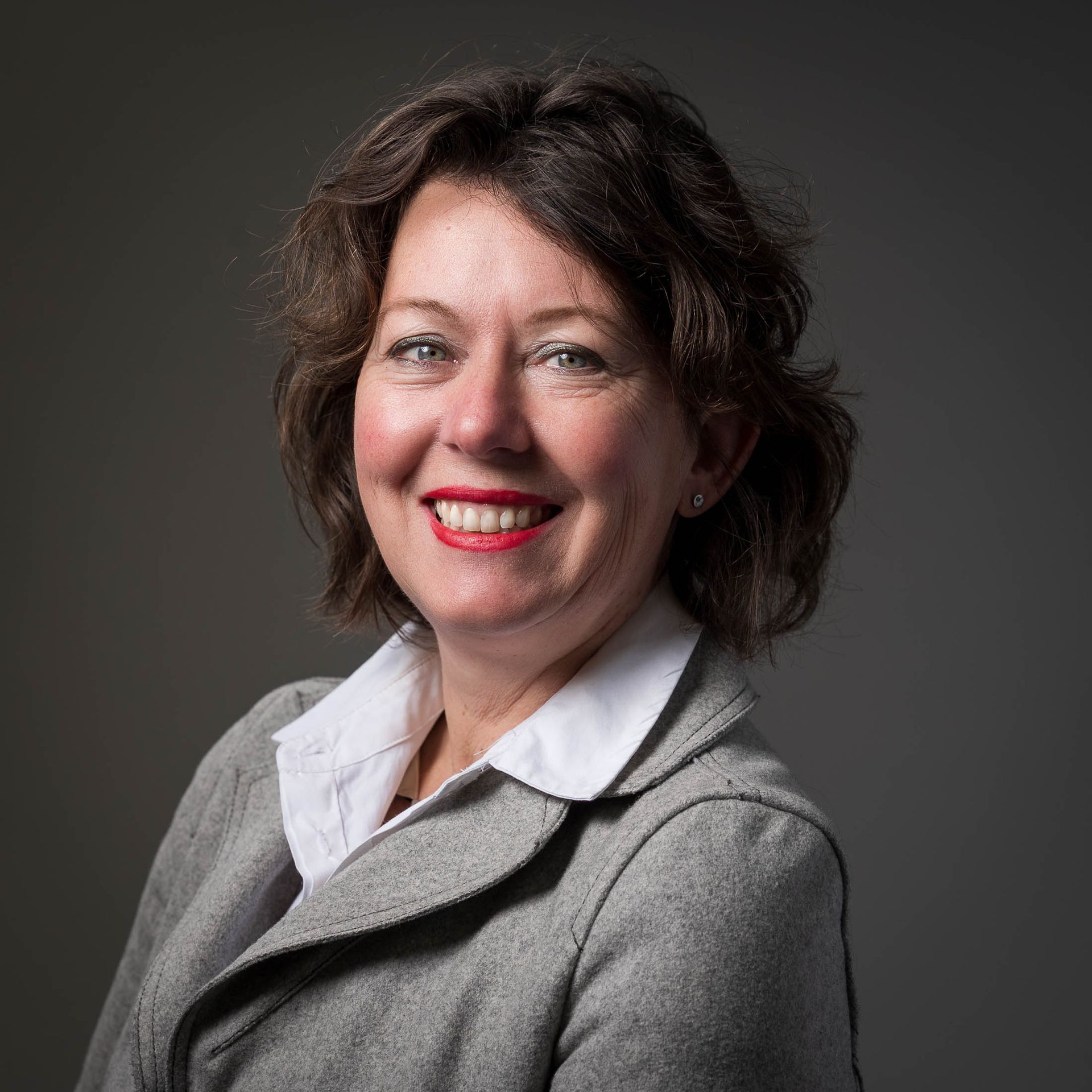 Een vrouw in een grijze jas en een wit shirt lacht naar de camera.