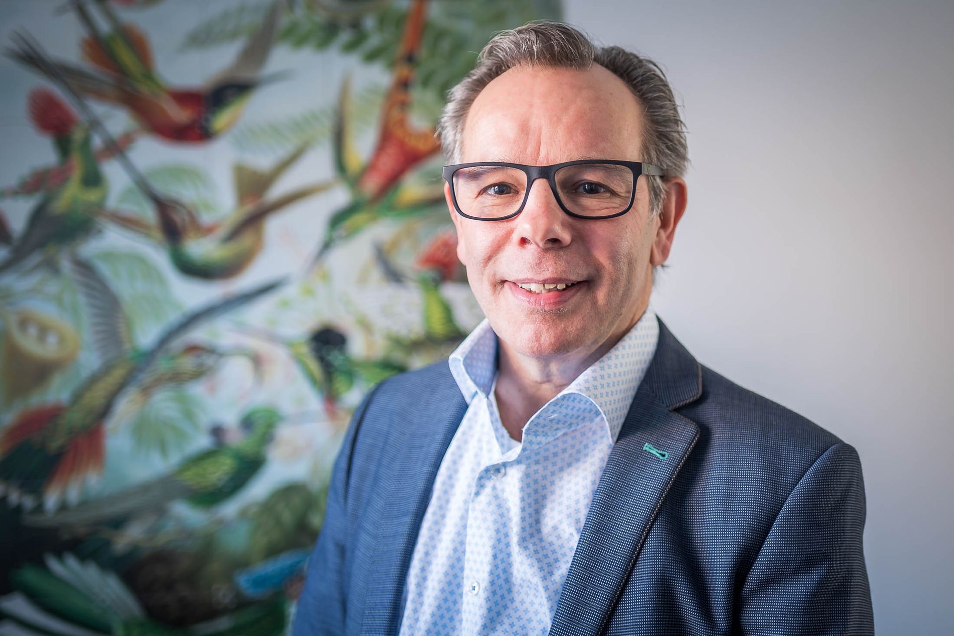 Een man in pak en bril staat voor een schilderij van vogels.