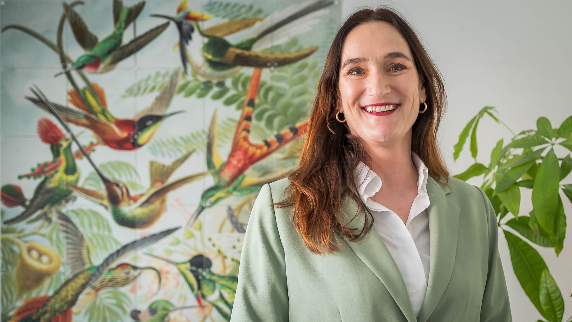 Een vrouw in een groen jasje staat voor een schilderij van vogels.