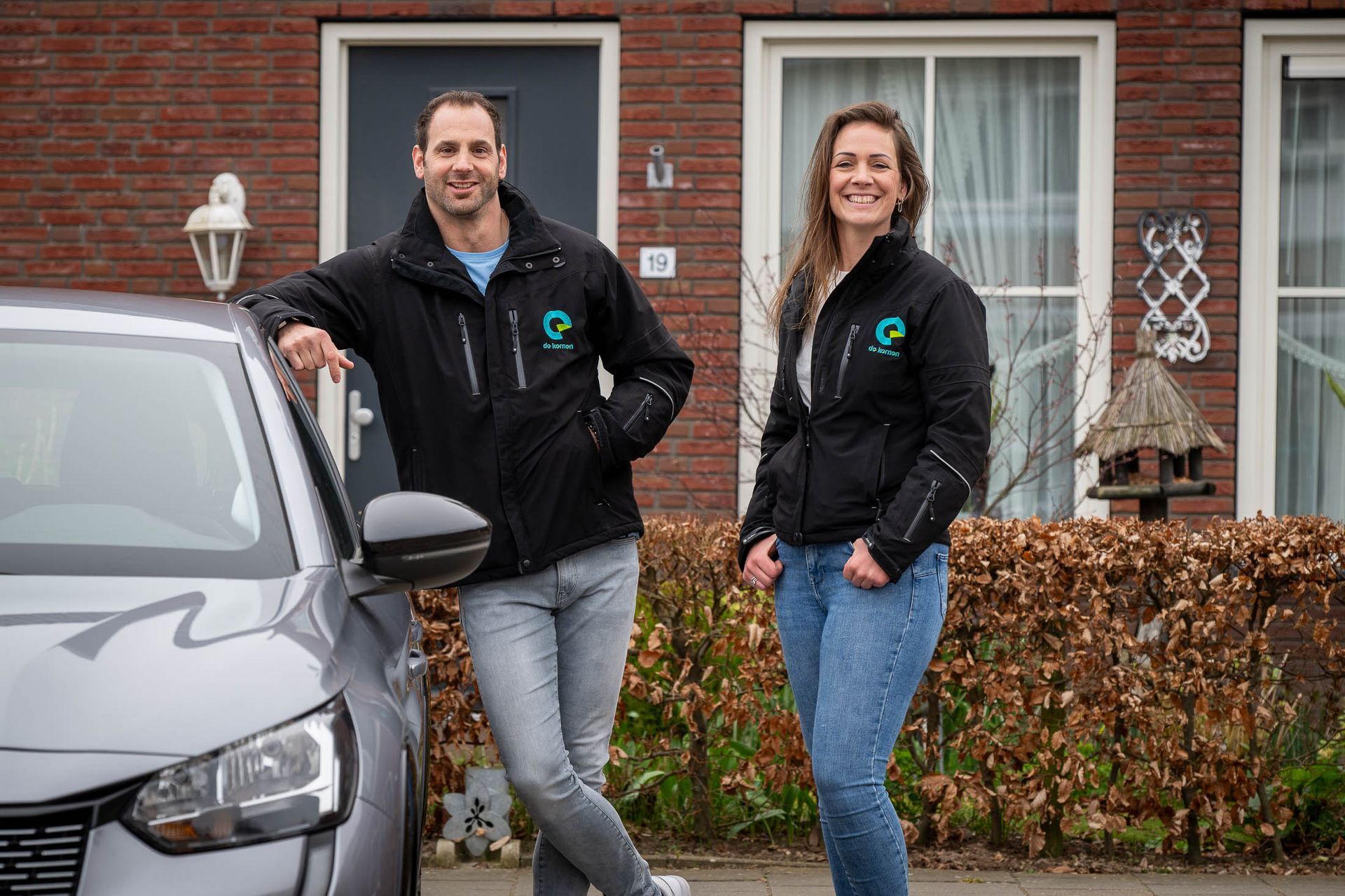 Een man en een vrouw staan naast een auto voor een bakstenen huis.