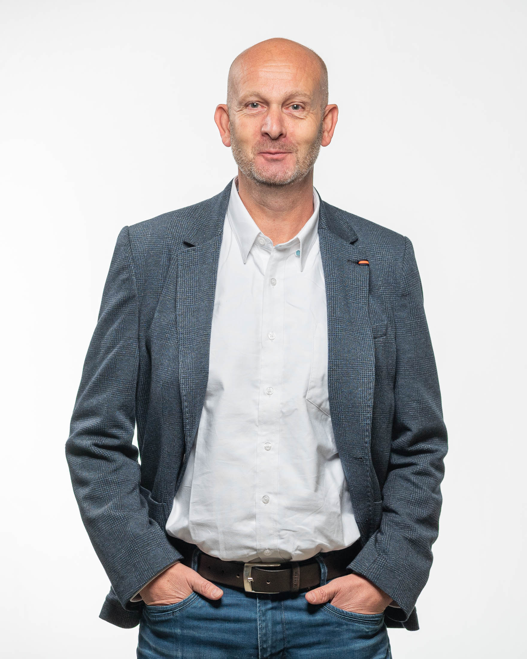 Een kale man draagt een blauw jasje en een wit overhemd en staat met zijn handen in zijn zakken.