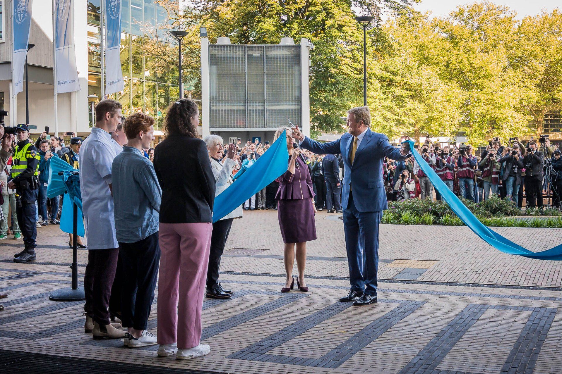 Een man in een pak houdt een blauw lint vast voor een groep mensen.