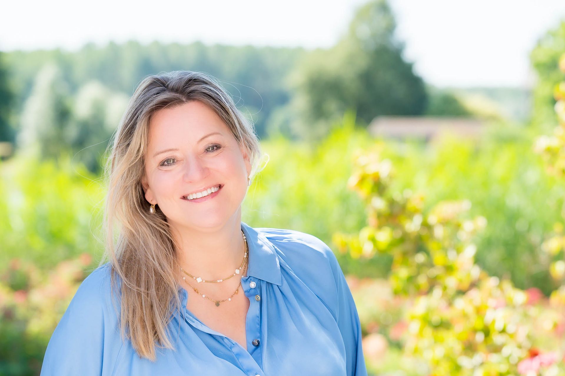 Een vrouw in een blauw shirt lacht voor een veld vol bloemen.