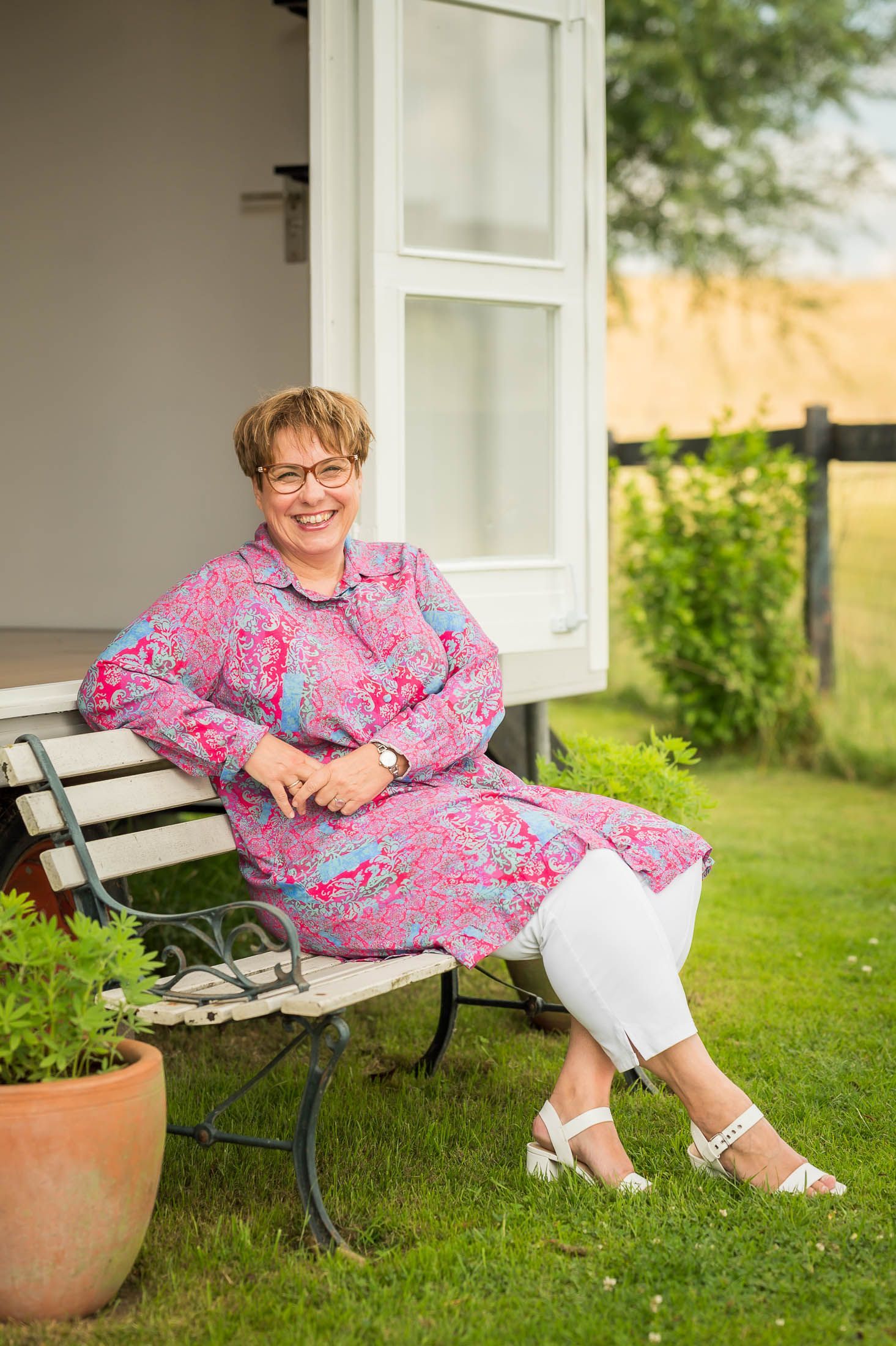 Een vrouw zit op een bankje in het gras naast een potplant.