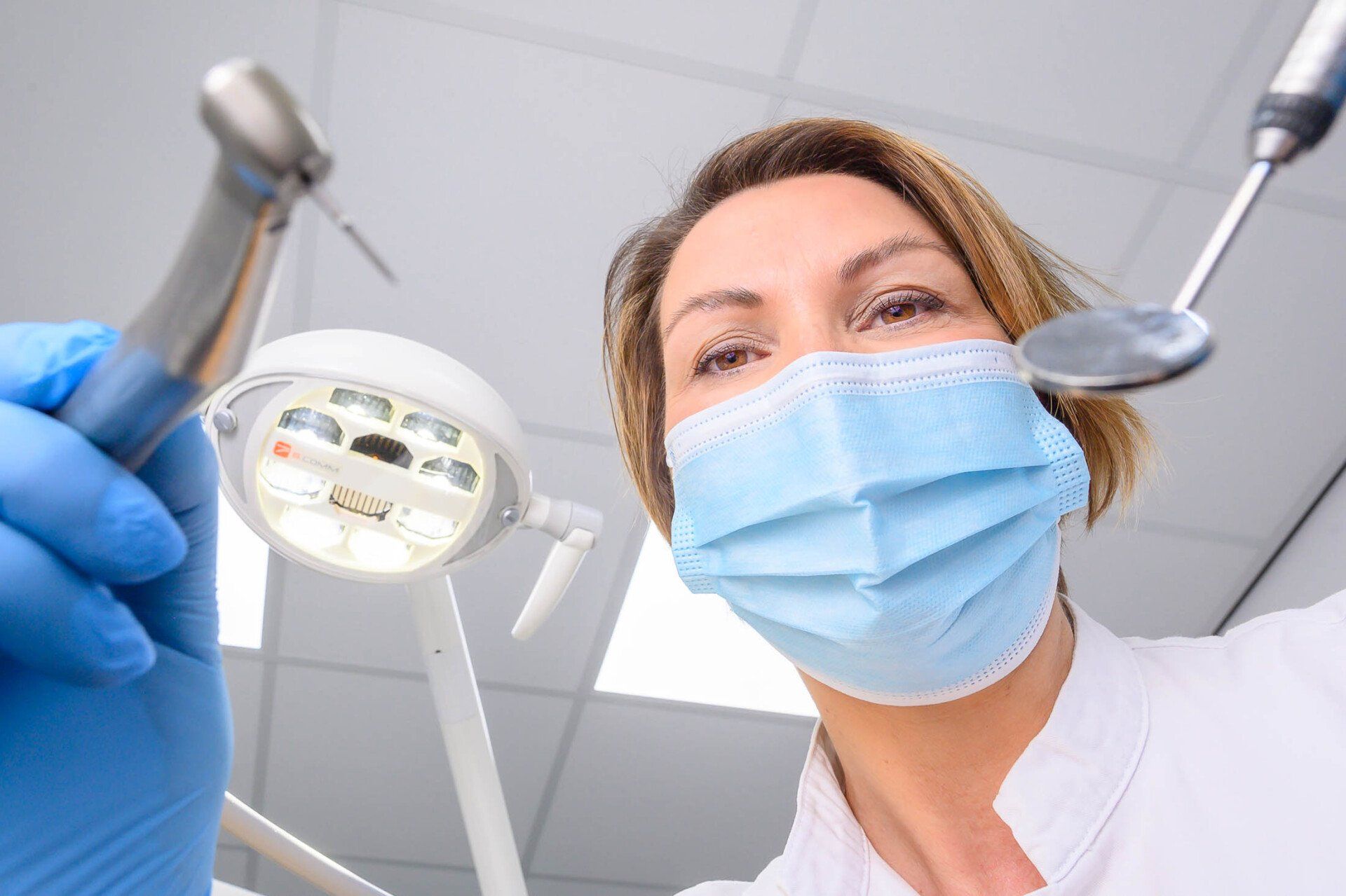 Een vrouwelijke tandarts draagt een masker en handschoenen en houdt een boor en een spiegel vast.