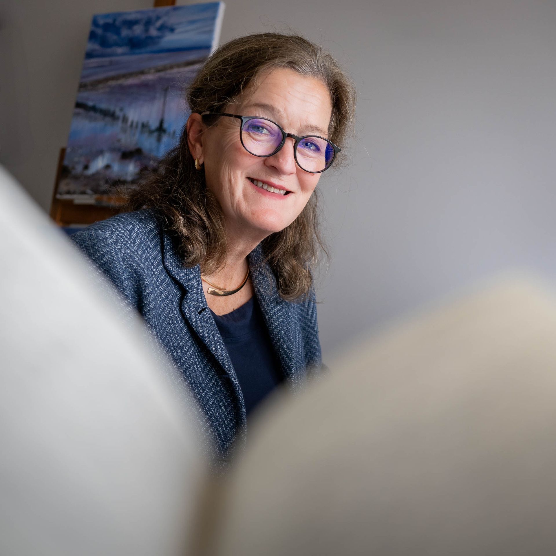 Een vrouw met een bril en een blauw jasje staat lachend voor een schilderij.