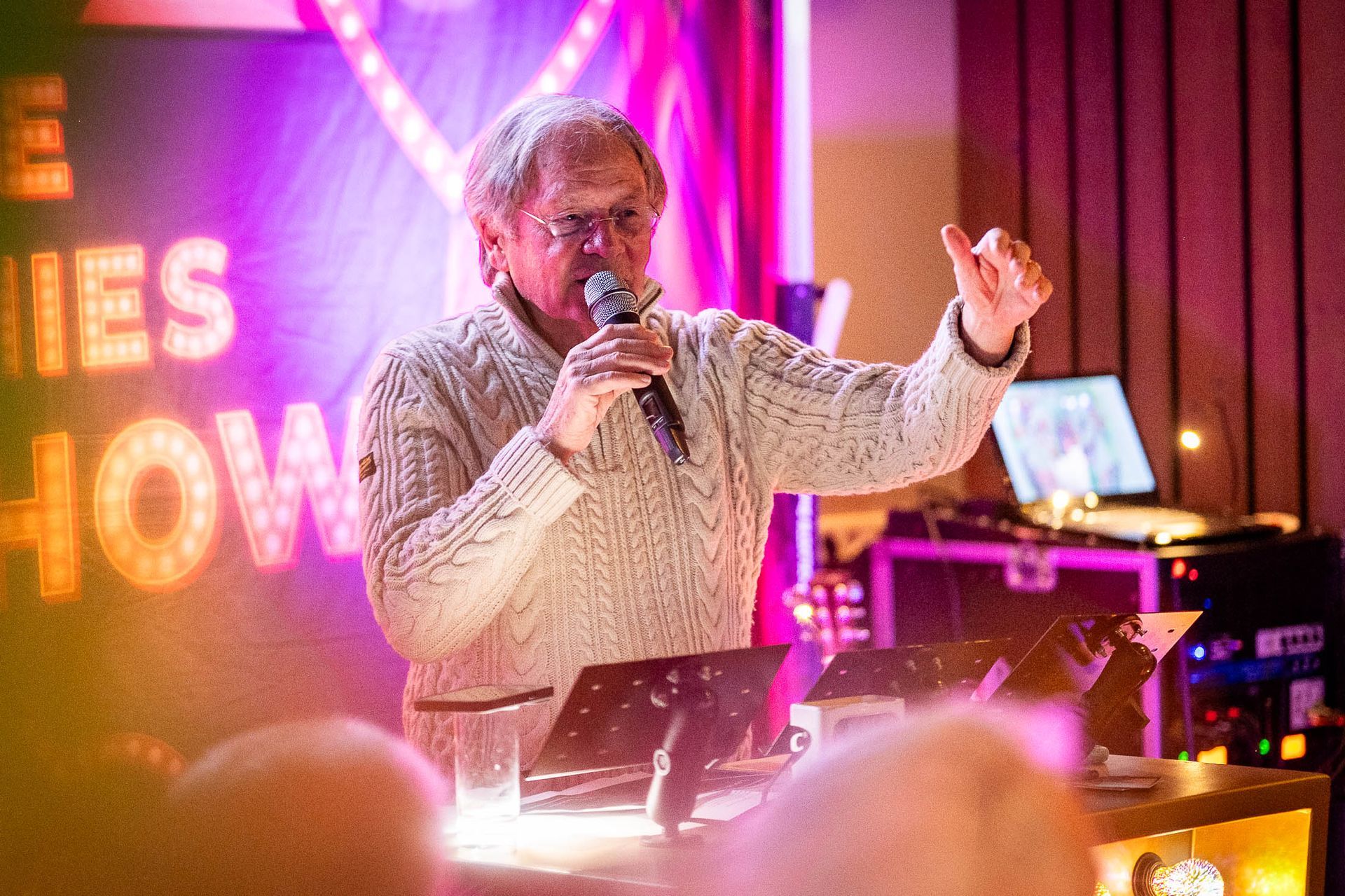 Man spreekt in een microfoon op het podium; opgeheven arm. Theatrale setting, kleurrijke belichting.