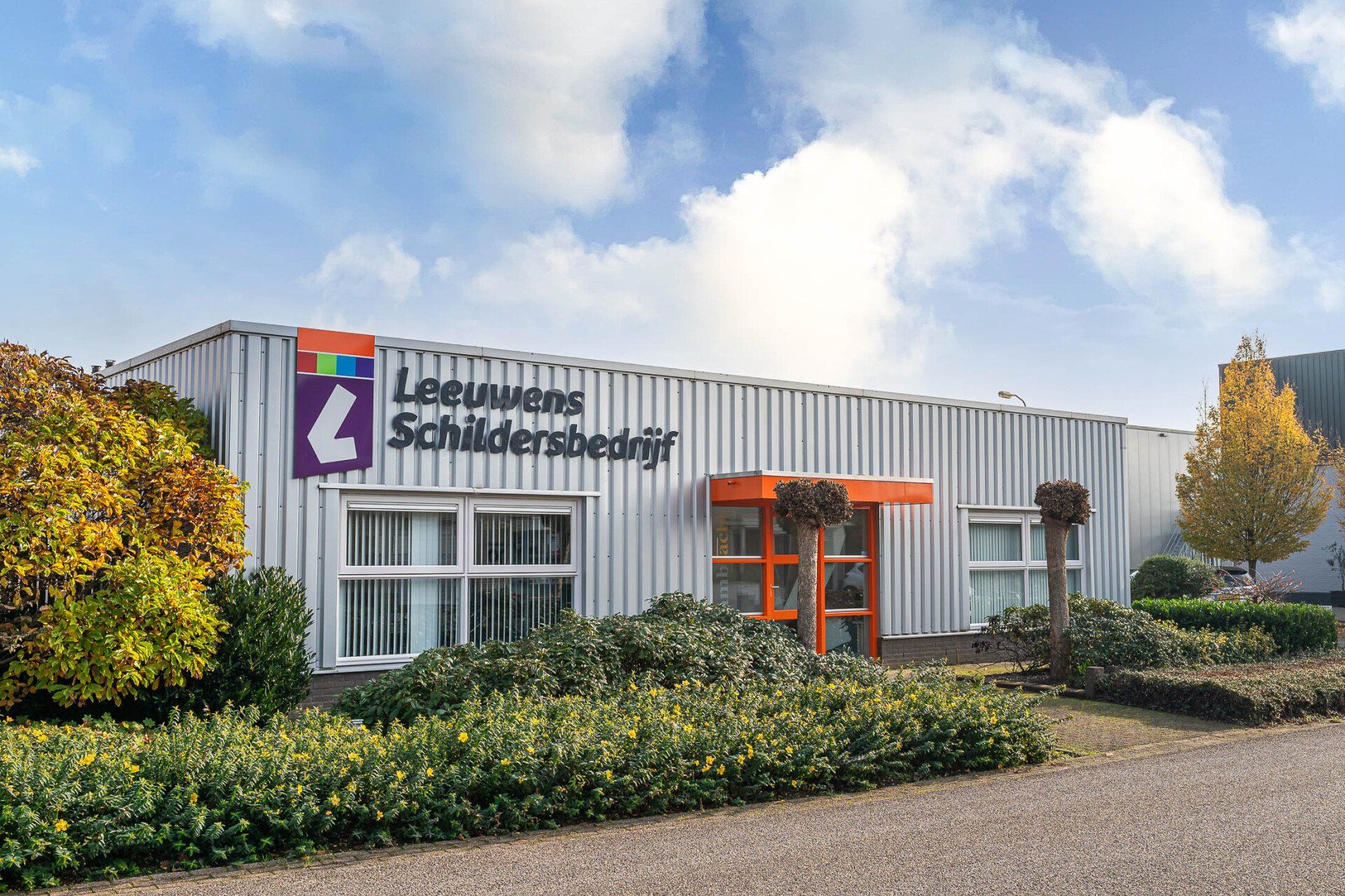 Een groot wit gebouw met een paars logo erop, omgeven door struiken en bomen.