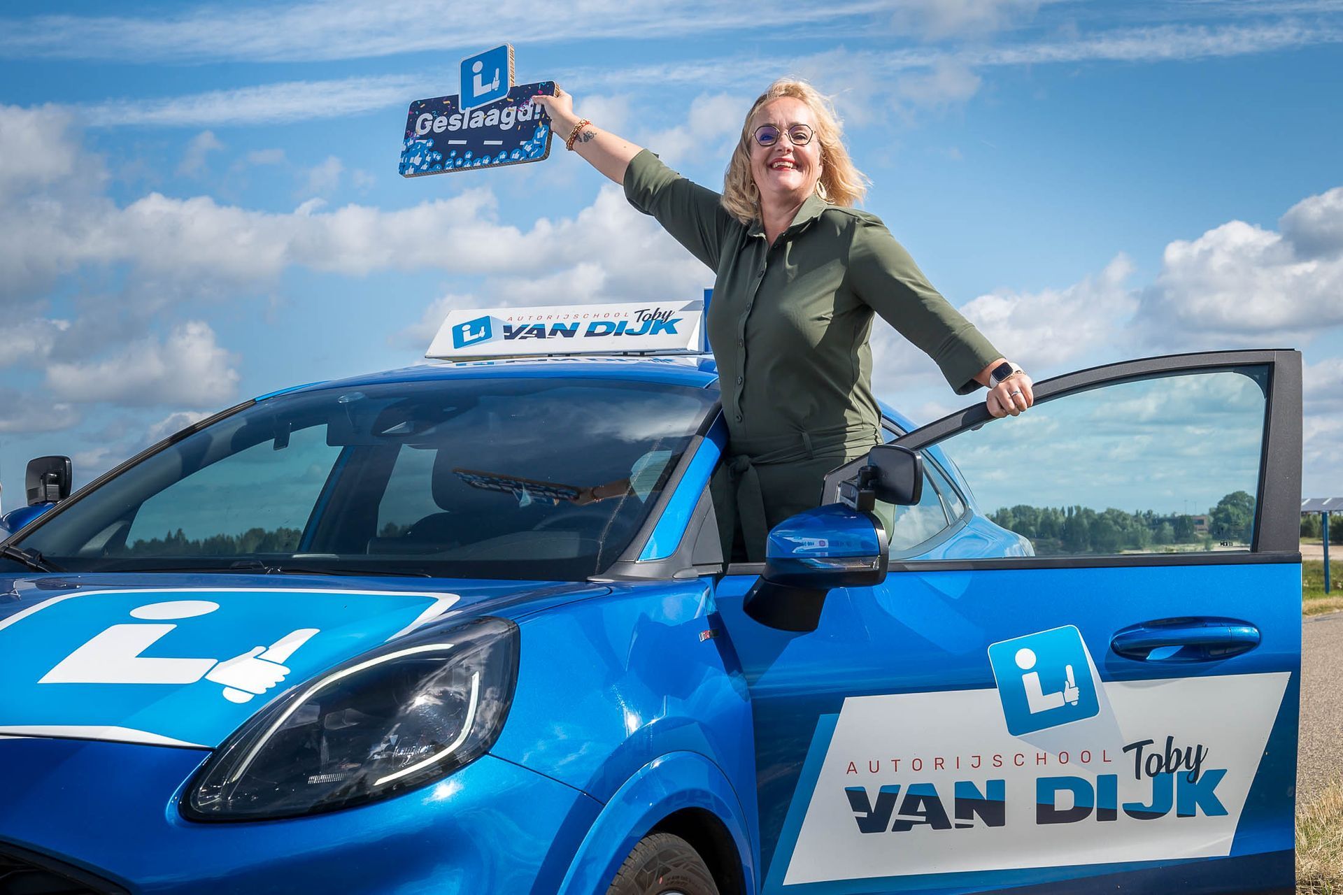 Een vrouw staat naast een blauwe Van Dijk auto en houdt een bord vast.