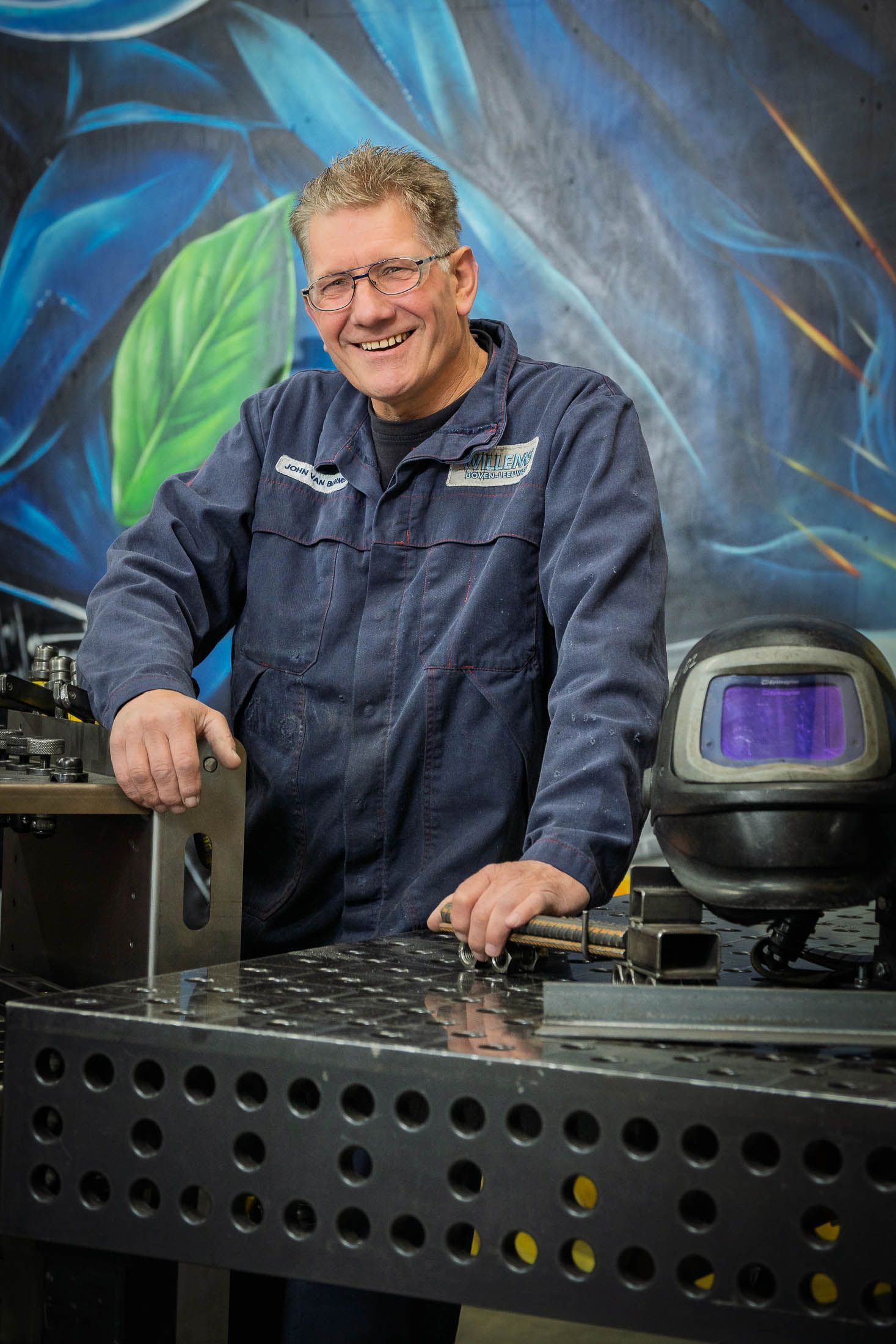 Een man in werkkleding glimlacht en leunt tegen een metalen tafel. Een lashelm staat ernaast, tegen de achtergrond van een blauwe muurschildering.