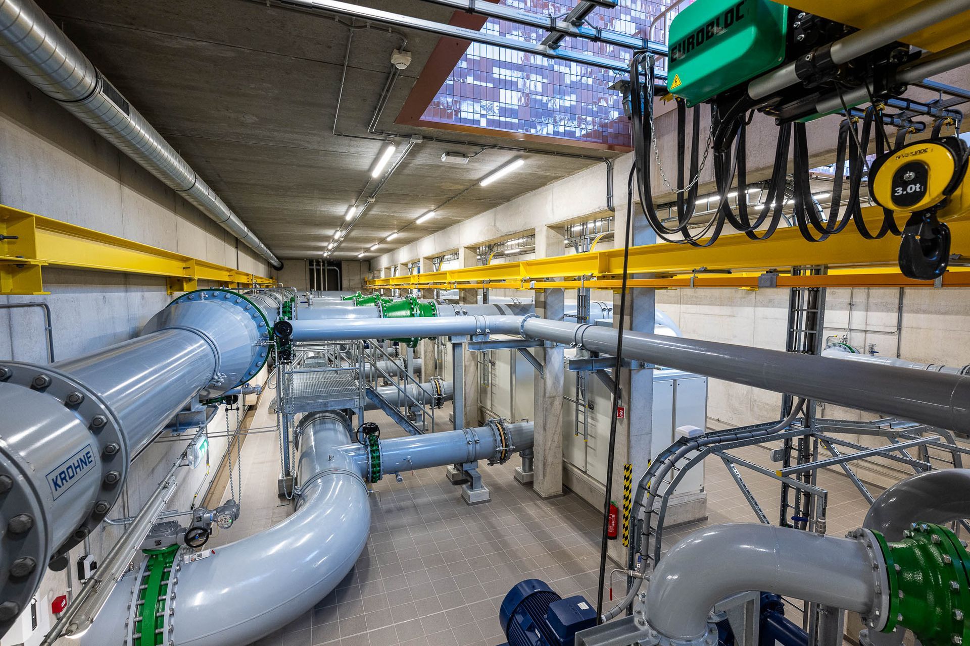 Industrieel interieur met grote pijpen en machines, gele steunbalken en een bovenloopkraan.