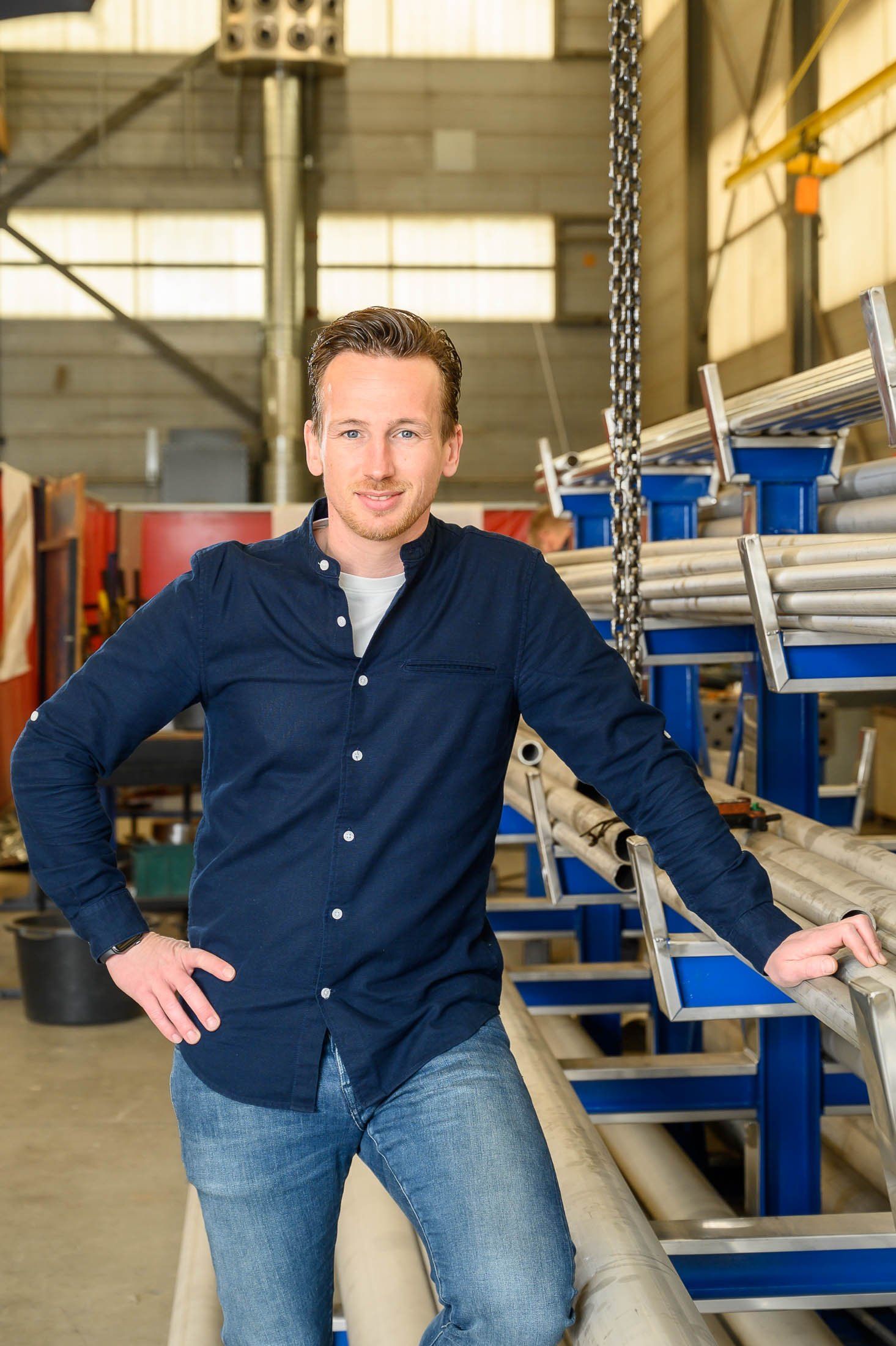 Een man in een blauw overhemd en spijkerbroek staat in een pakhuis met zijn handen in zijn zij.