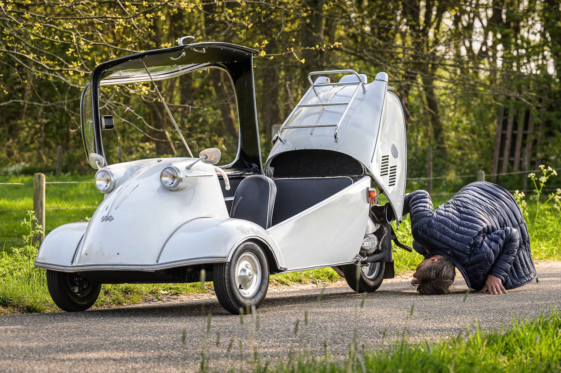 Een man knielt voor een witte auto met de motorkap omhoog.