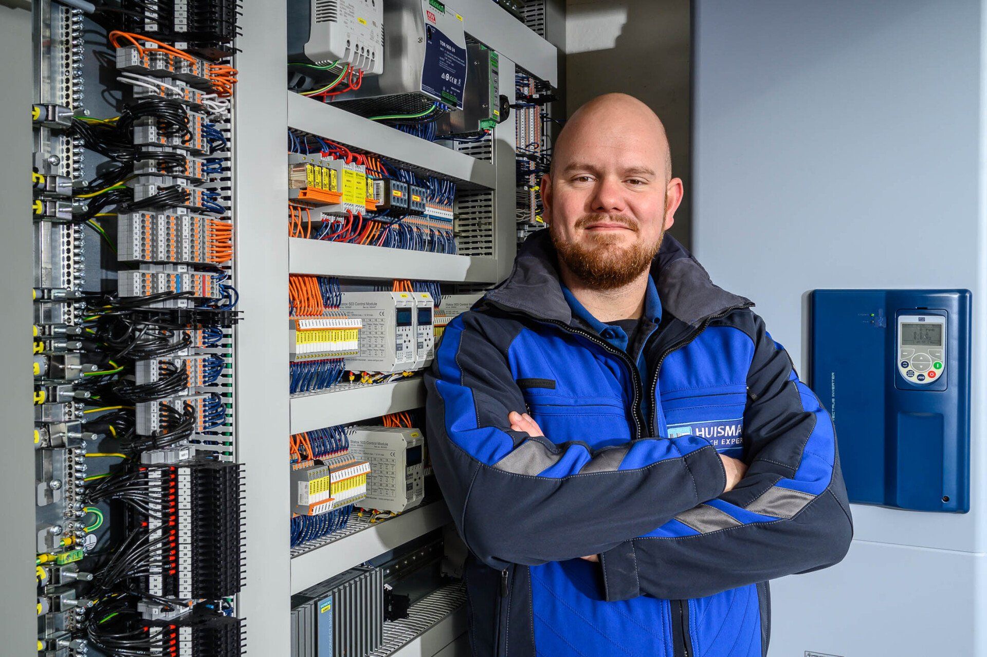 Een man met een baard staat voor een elektriciteitskast, met zijn armen over elkaar.
