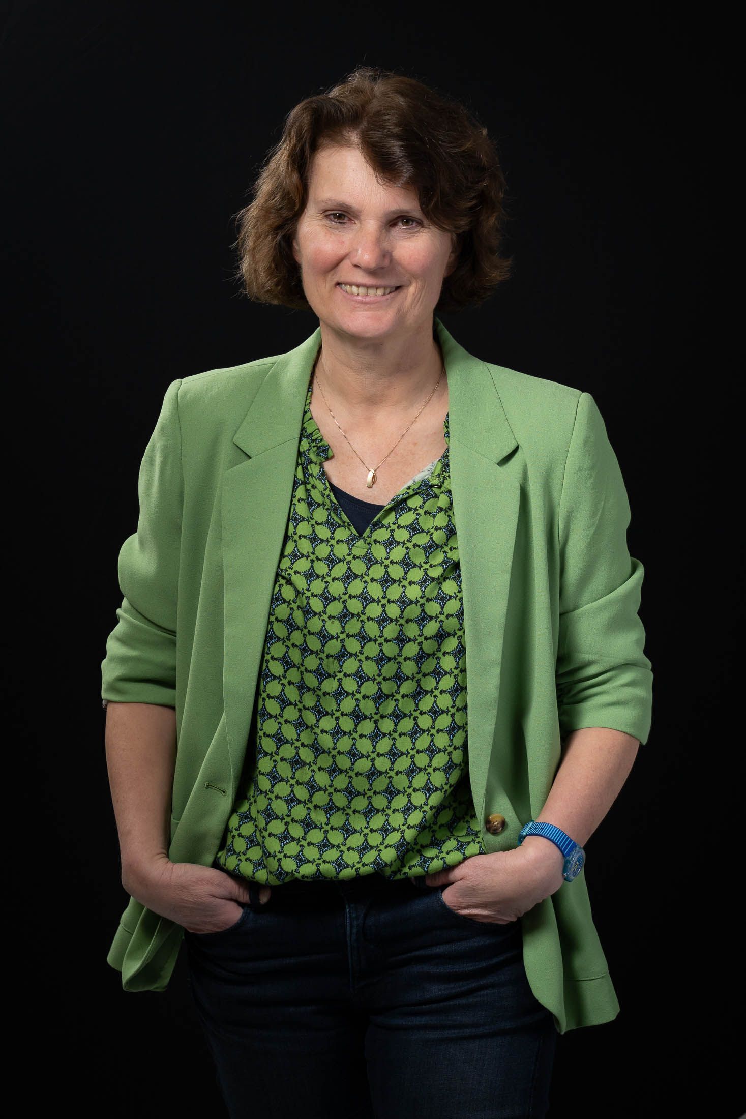 Een vrouw draagt een groen jasje en een groen shirt en staat met haar handen in haar zakken.