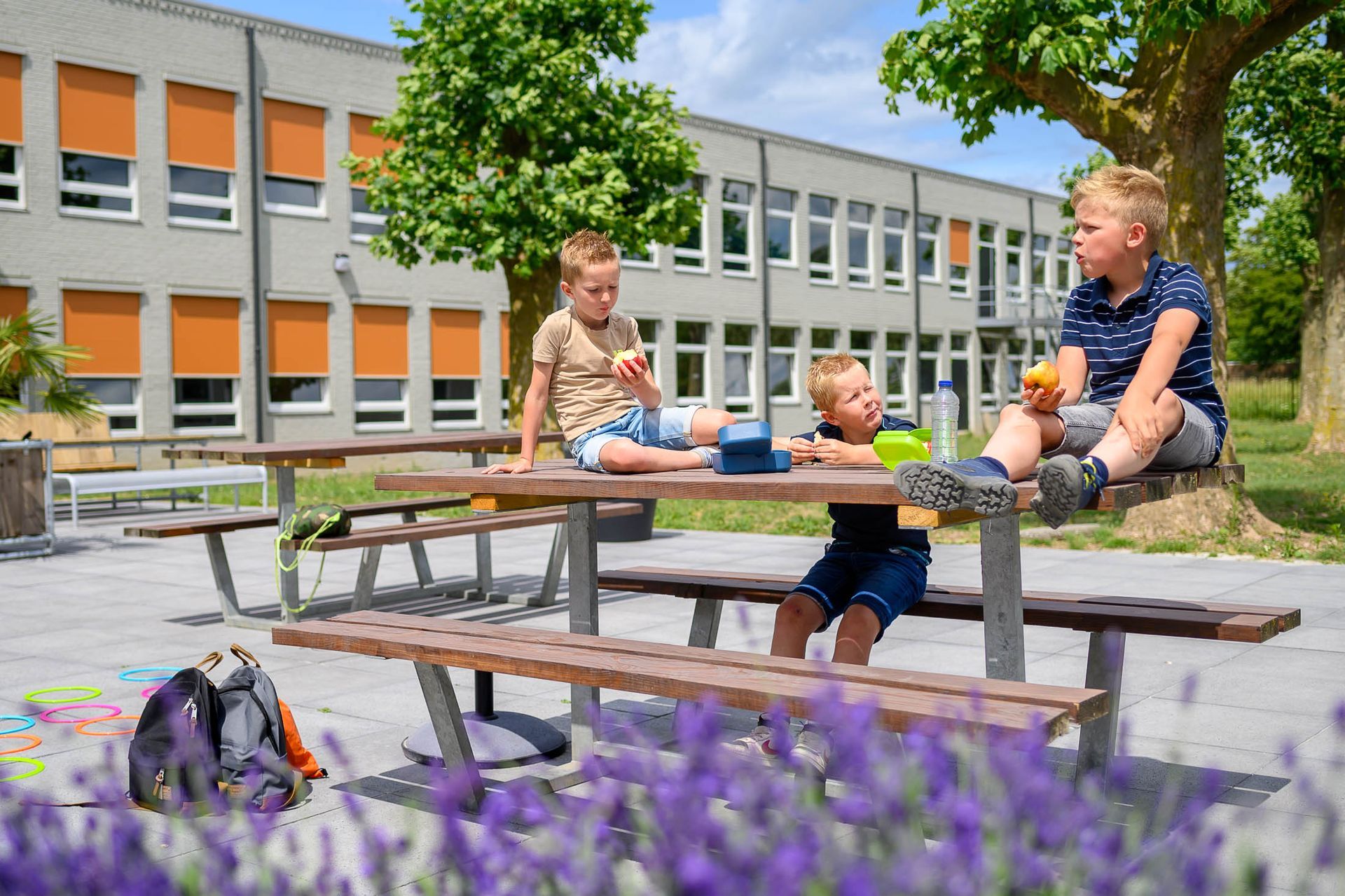 Drie kinderen zitten aan een picknicktafel voor een schoolgebouw.