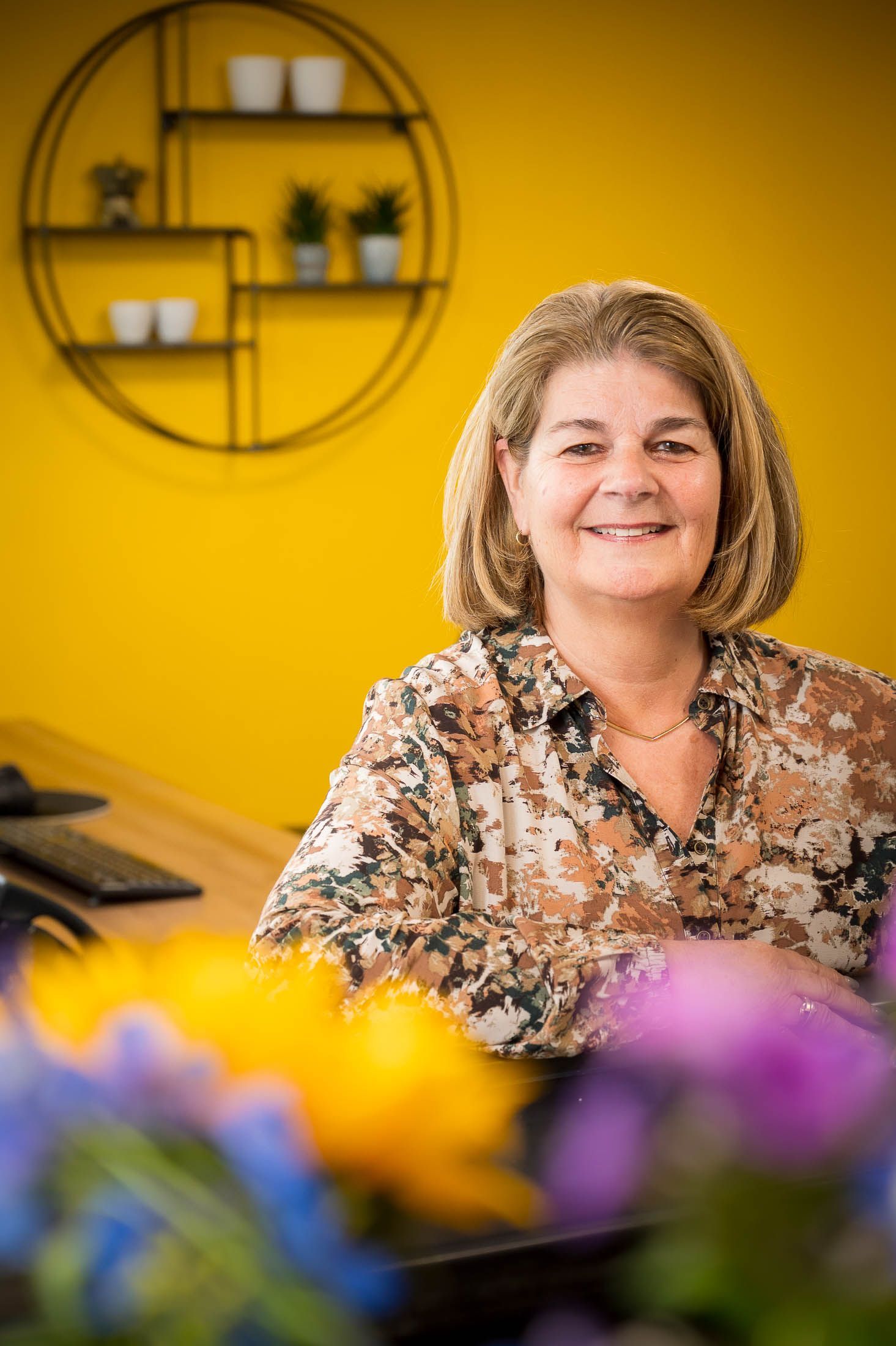 Een vrouw zit aan een bureau voor een gele muur met bloemen op de voorgrond.