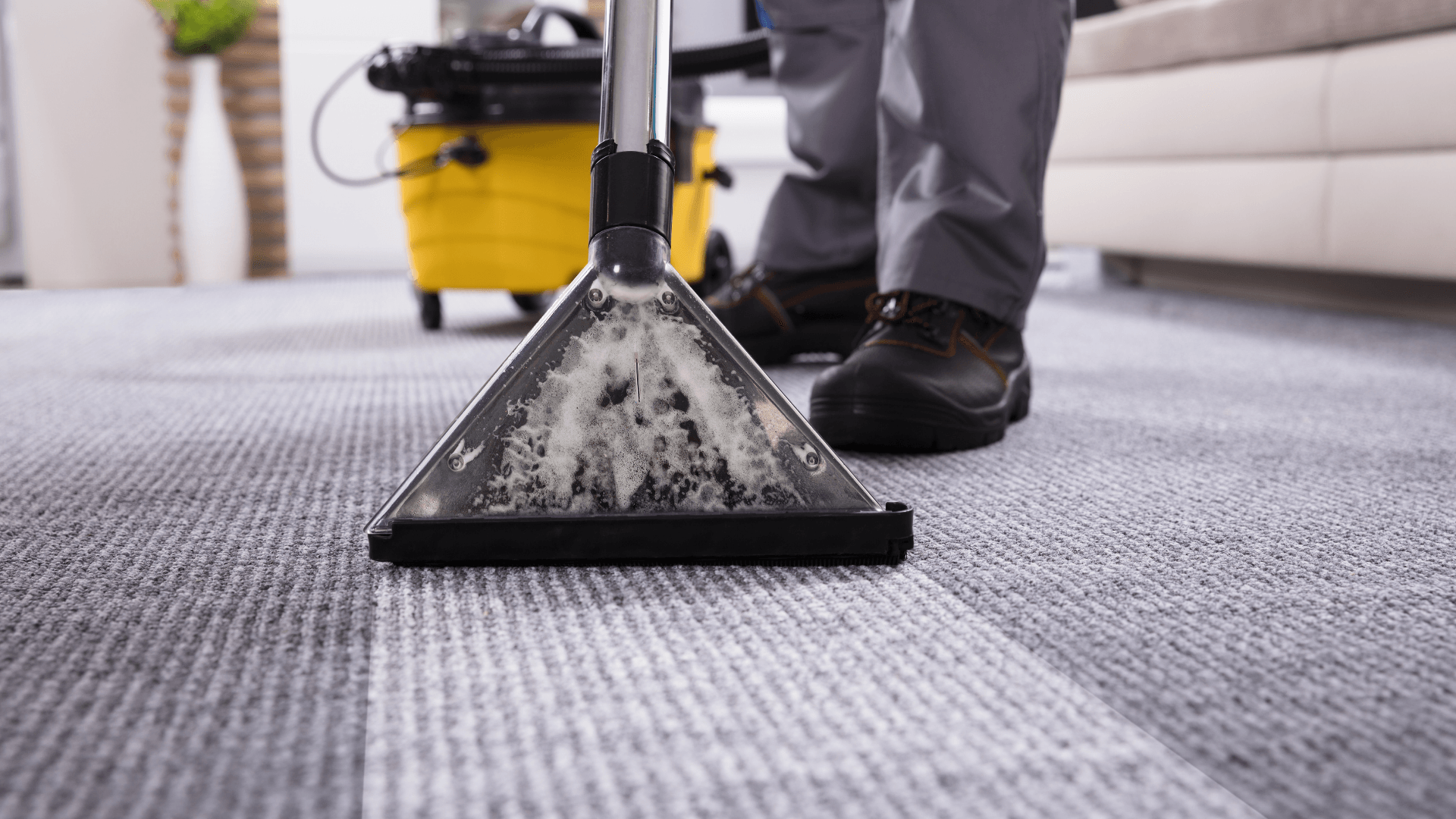 Carpet cleaner extracting dirt from a gray rug in a room, with a yellow bucket in the background