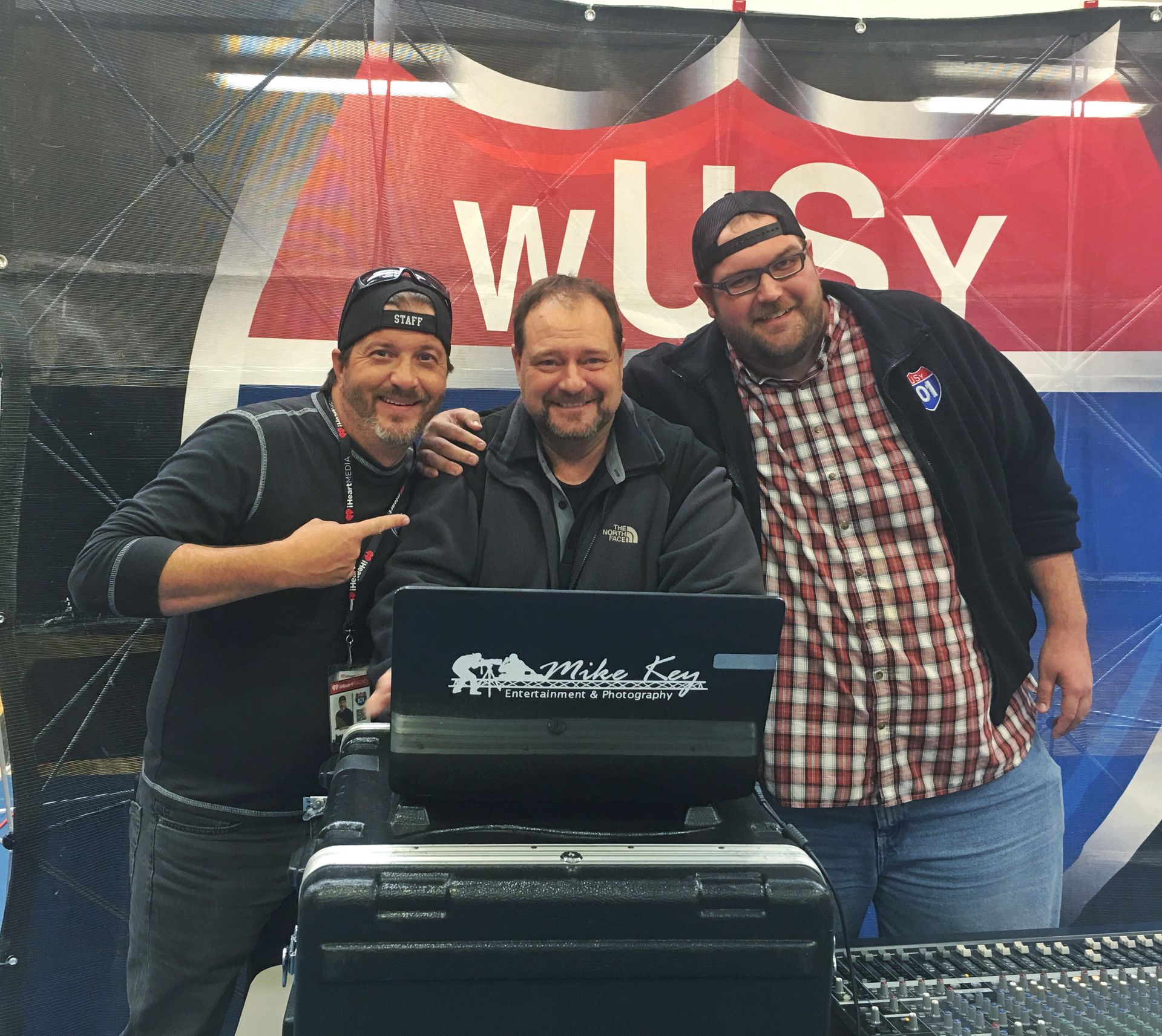 Three men pose behind a mixing console in front of a wUSy branded backdrop.
