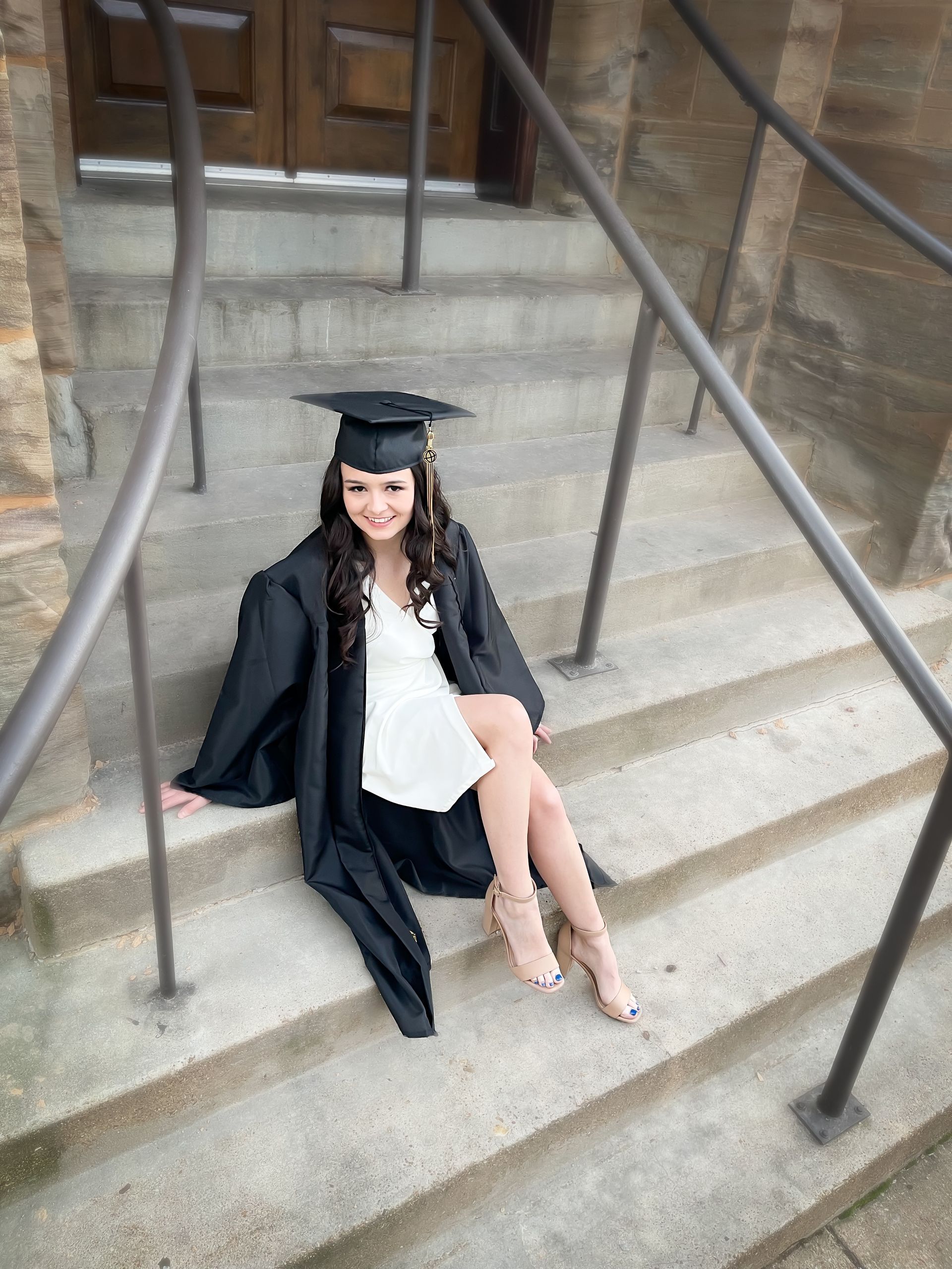 A person in a graduation cap and gown sits on stone steps in front of a wooden door, smiling at the camera.