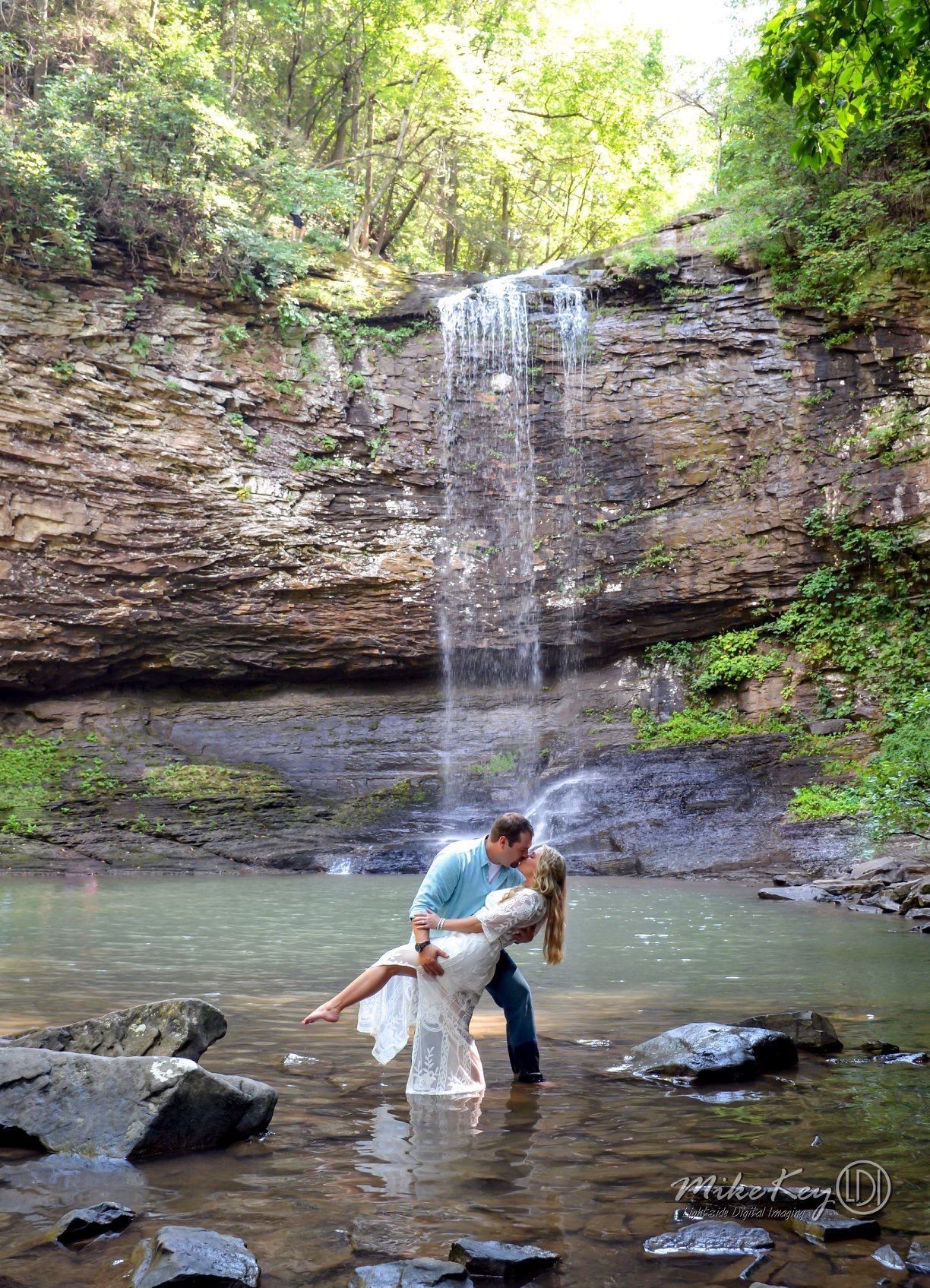 A couple embraces in a deep dip while standing in a shallow stream at the base of a waterfall surrounded by forest.